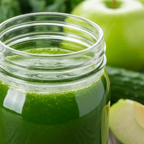 A green Happy Juice in a mason jar with fresh kale, cucumber, green apple, and lemon on a kitchen counter.