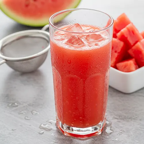 A glass of cold watermelon juice next to a strainer with leftover pulp, emphasizing freshness and texture.