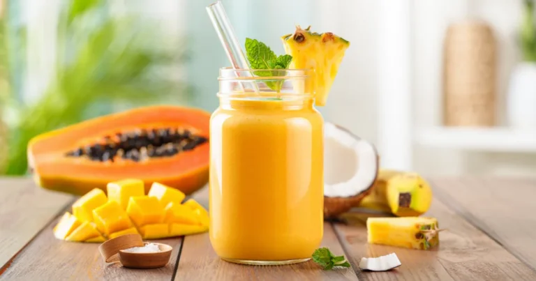 A glass jar filled with tropical smoothie surrounded by fresh mango, papaya, banana, and coconut on a wooden table.