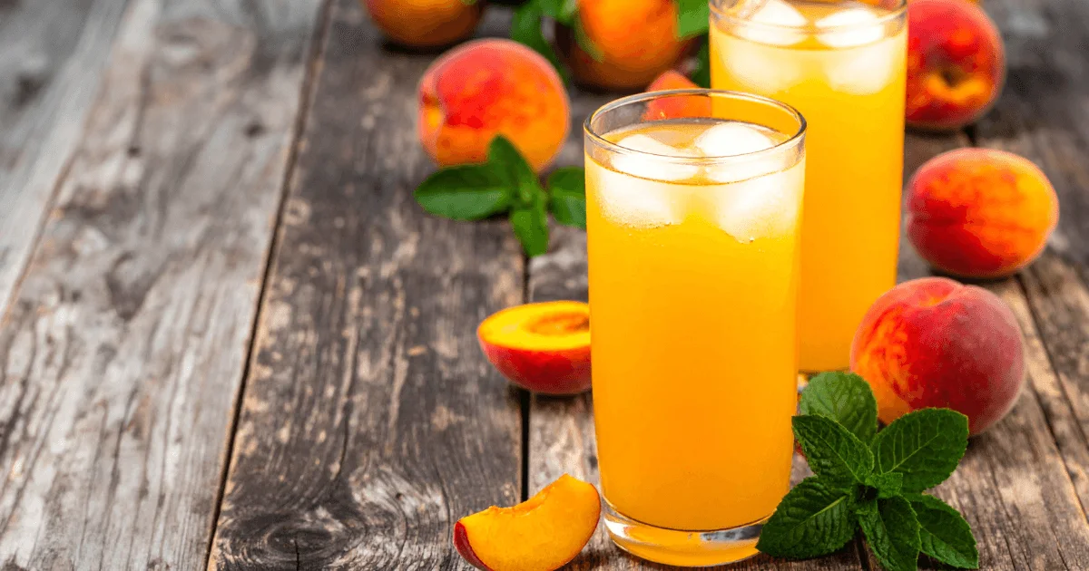 A tall glass of peach juice with ice, surrounded by fresh ripe peaches on a rustic wooden table, perfect for a refreshing summer drink.