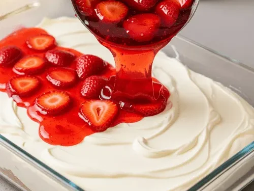 Strawberry Jell-O mixture with sliced strawberries being poured over the creamy layer of strawberry pretzel dessert.