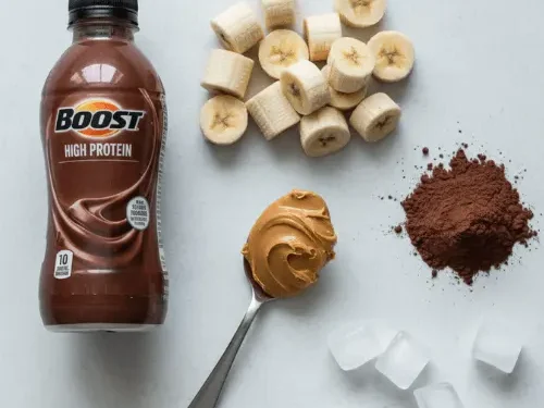 Fresh ingredients for a protein shake including a bottle of chocolate Boost, frozen bananas, peanut butter, and cocoa powder arranged on a counter.