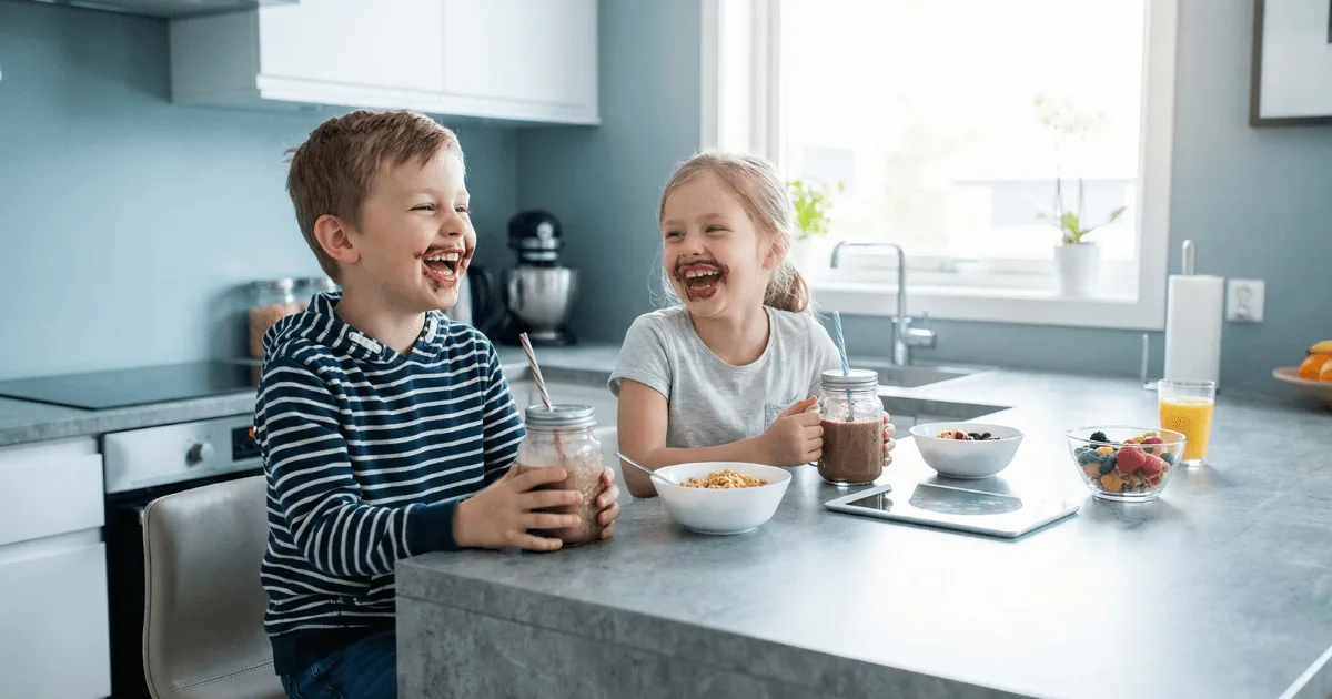 Two children happily drinking high-protein smoothies at a kitchen counter during a busy school morning.
