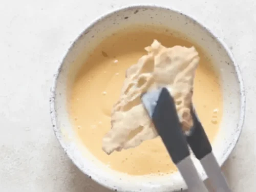 Croissant halves being dipped into egg custard for French toast preparation. The croissants absorb flavor while keeping their flaky structure.